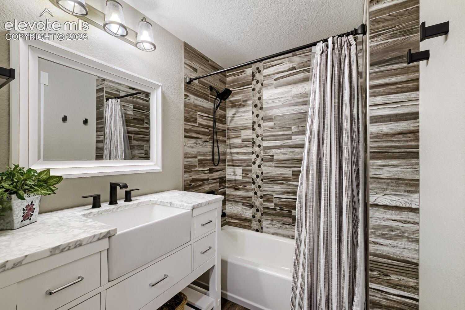 White vanity with a farmhouse sink, marble-look countertop, black hardware, and a tiled shower surround with accent strip