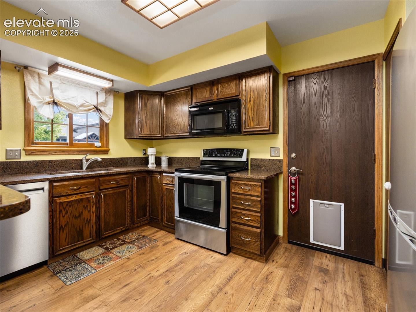 Wood-finish flooring, dark wood cabinetry, stainless steel appliances, dark stone-finish countertops, and a window with natural light