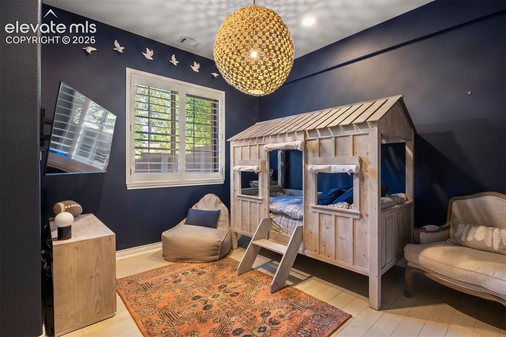 Bedroom featuring a built-in house-frame bed, light wood-finish flooring, a window with white louvered shutters, and a woven spherical pendant light fixture