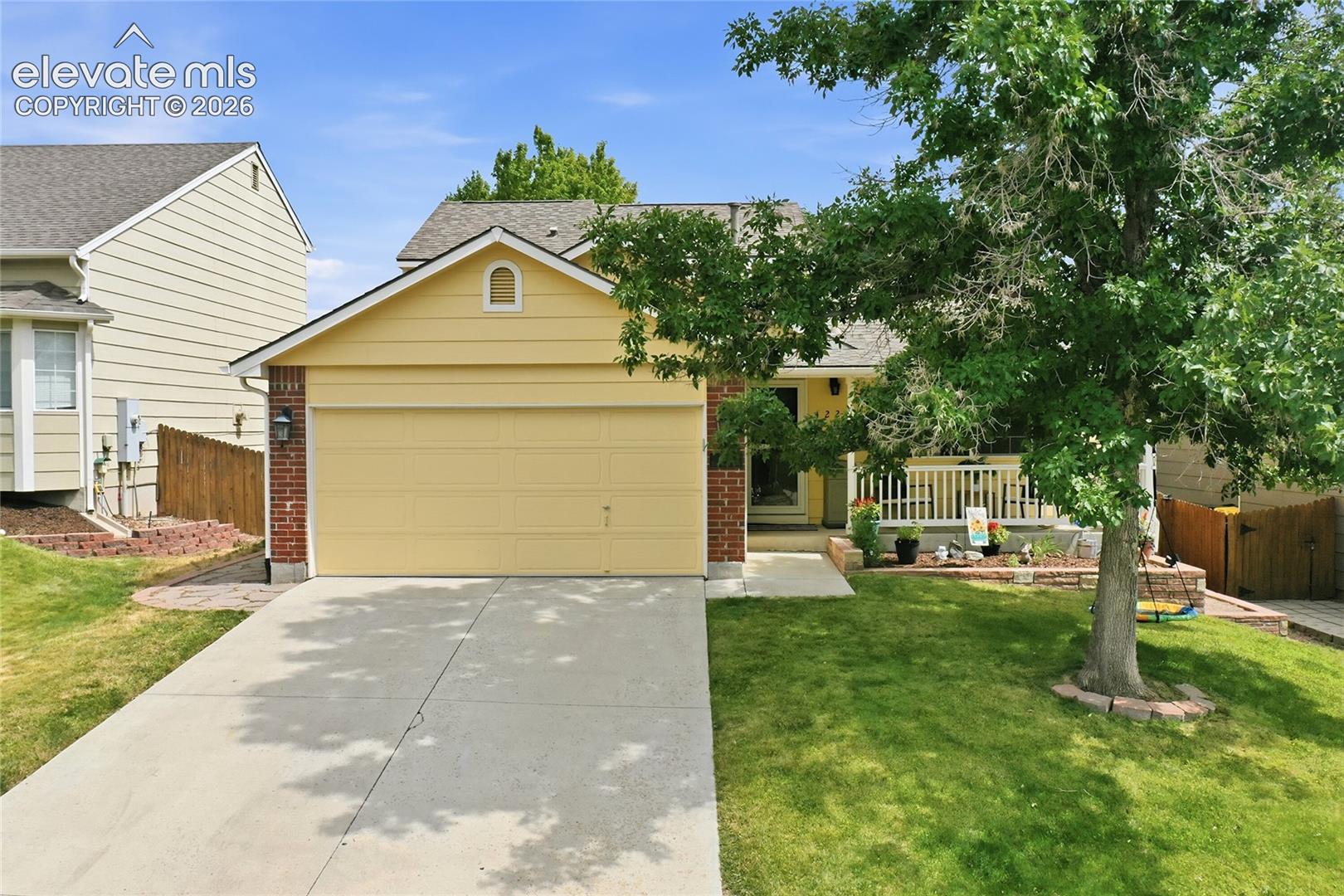 Single-story exterior featuring a yellow lap siding façade, brick accents, an attached two-car garage, and a covered front porch with a wood-finish railing