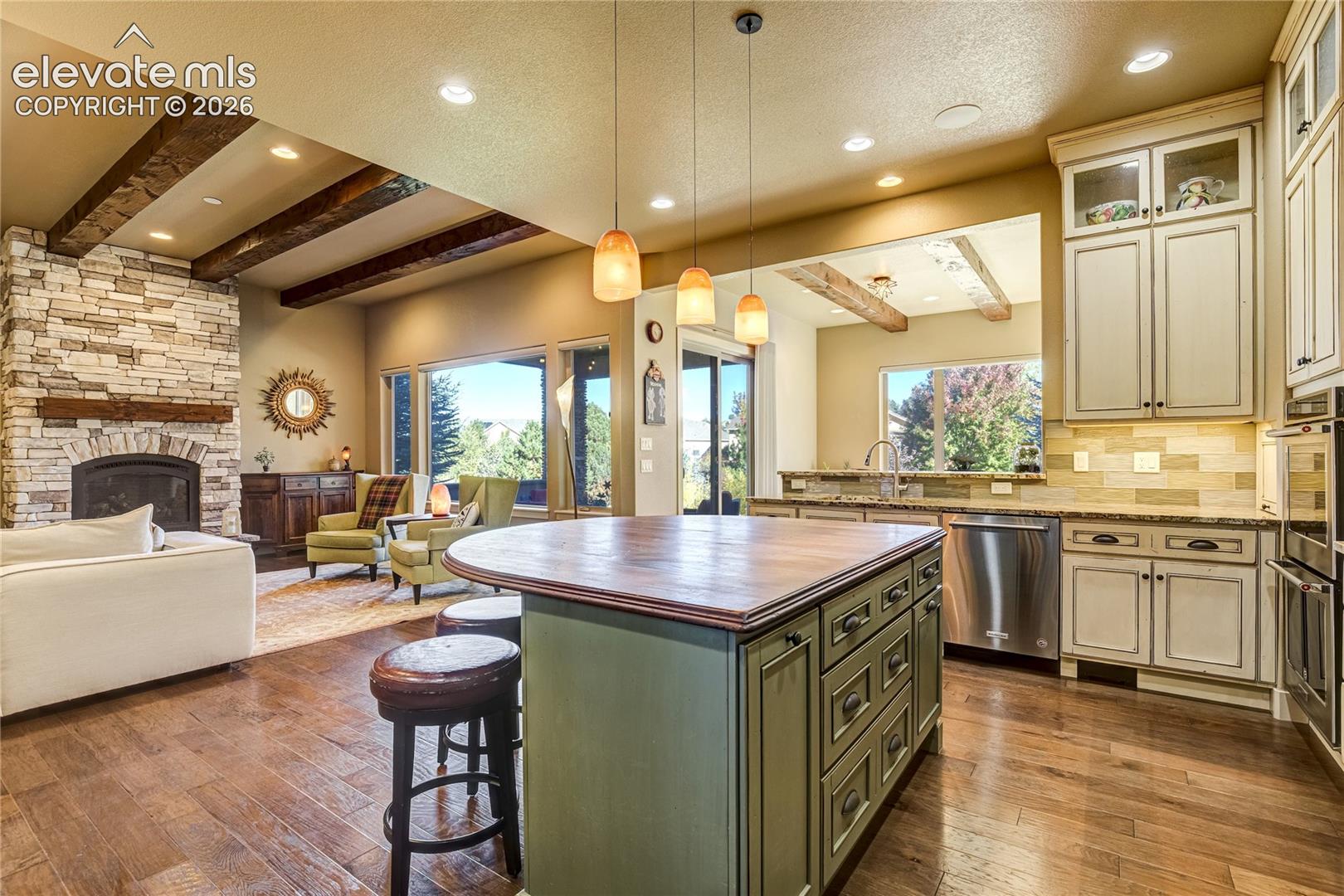 The butcher-block island offers a warm contrast to the granite perimeter counters, while pendant lighting + rich wood flooring bring character to the kitchen. Open sightlines toward the stone fireplace make entertaining feel effortless.