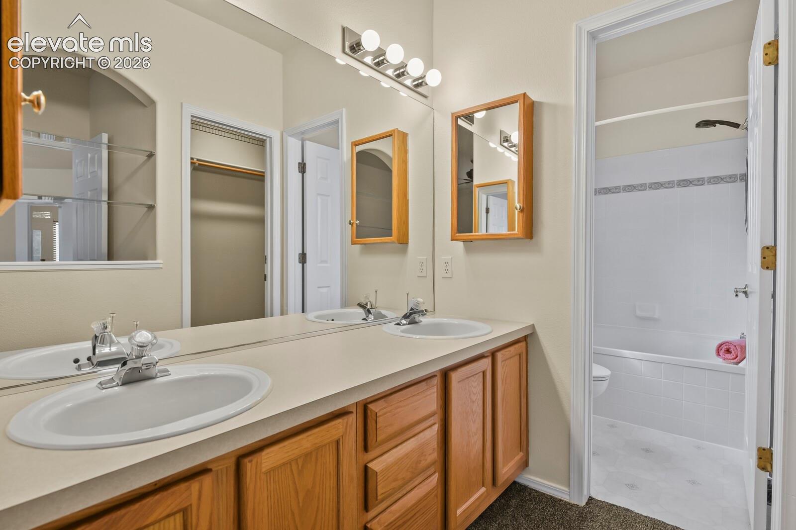 Dual-sink vanity with wood-finish cabinetry, solid-surface countertop, full-width mirror, and two wall-mounted medicine cabinets, plus a separate bathing area featuring a tile-surround tub