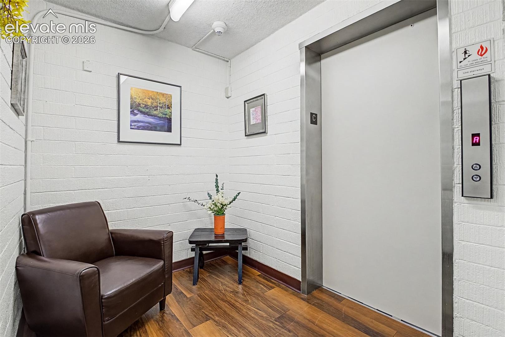 Elevator access alongside a waiting area featuring wood-finish flooring, white brick walls, and overhead fluorescent lighting