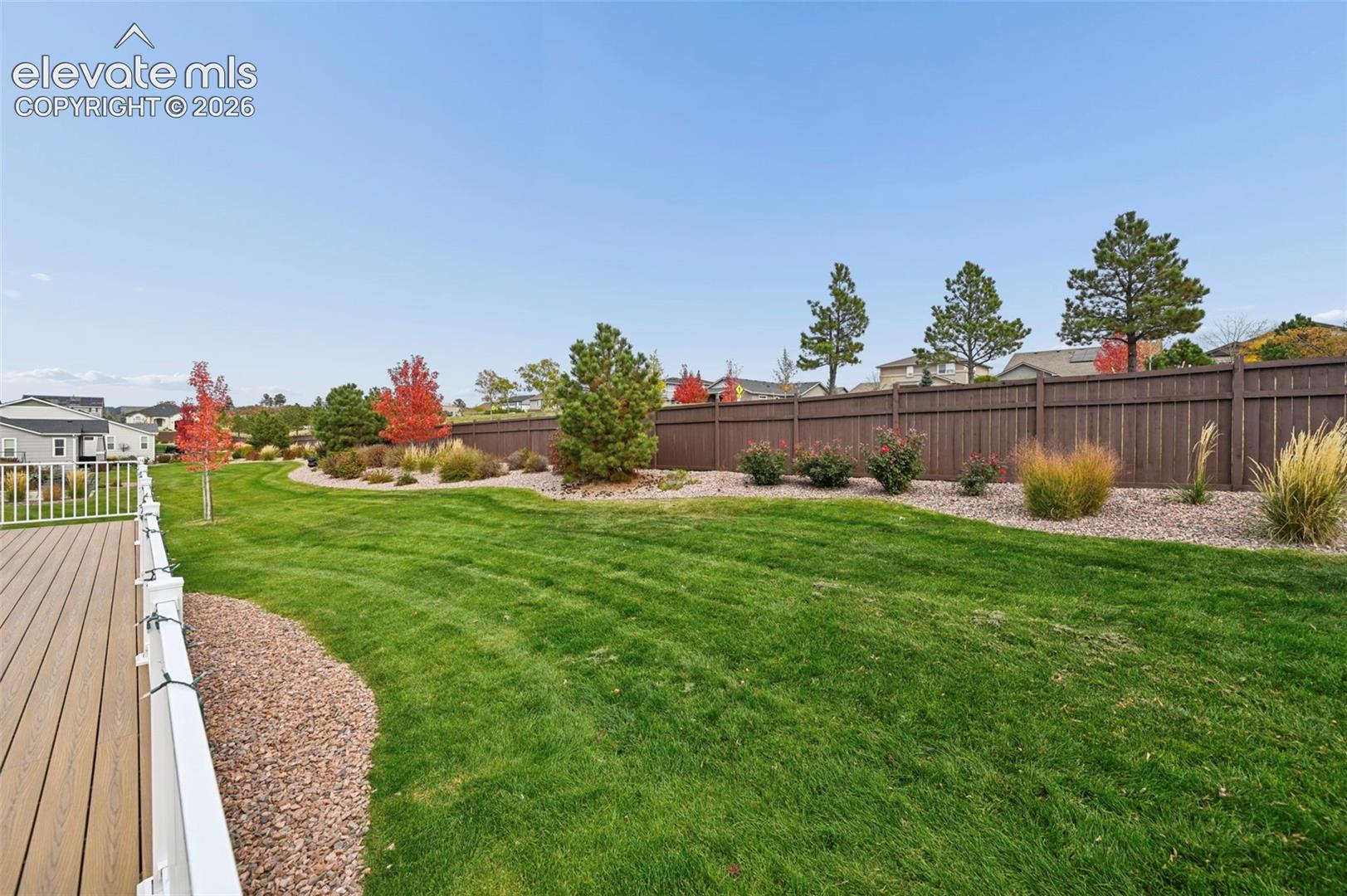 Rear deck featuring composite planking and white railings, overlooking an expansive lawn with mature trees, established perennial landscaping, and a privacy fence