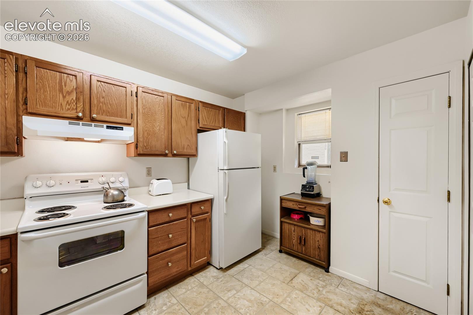 Kitchen featuring wood cabinetry, white appliances, durable tile flooring, and a window for natural light