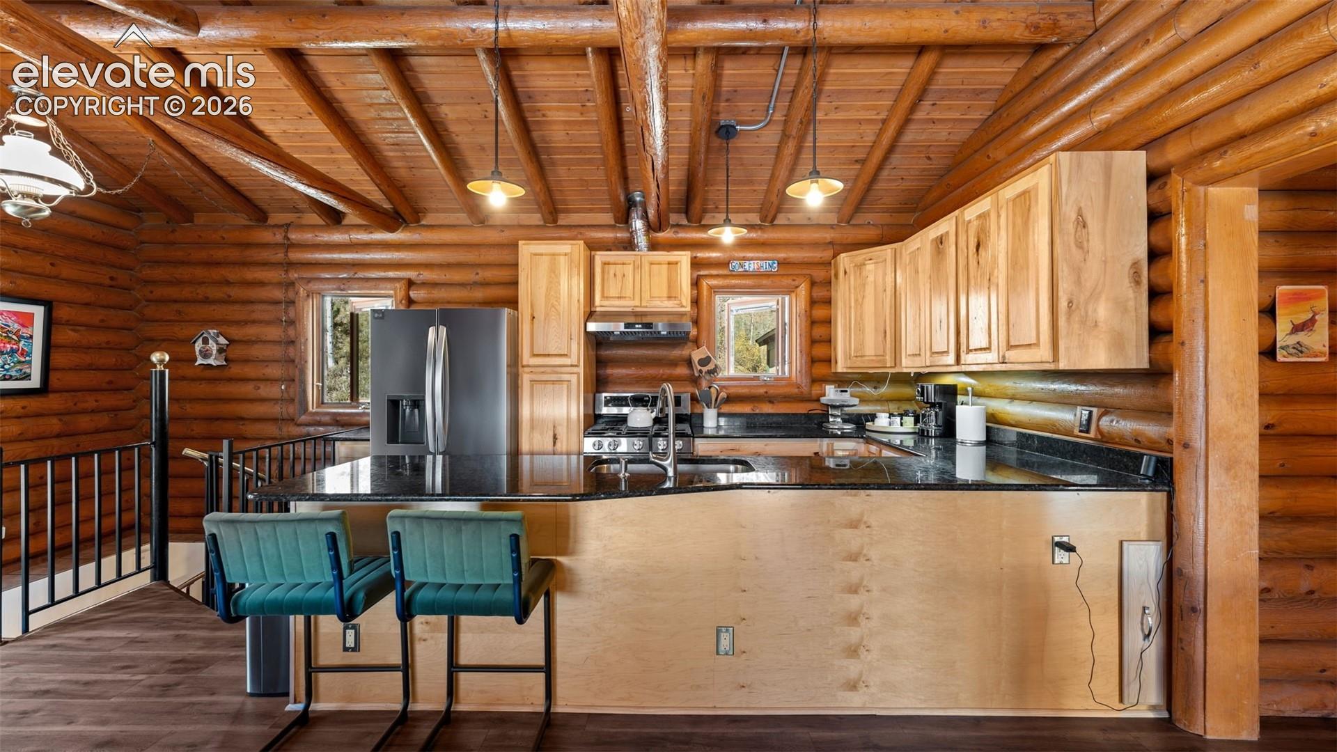 Rustic kitchen featuring exposed log walls and beamed ceilings, dark stone countertops, natural wood cabinetry, and multiple pendant light fixtures