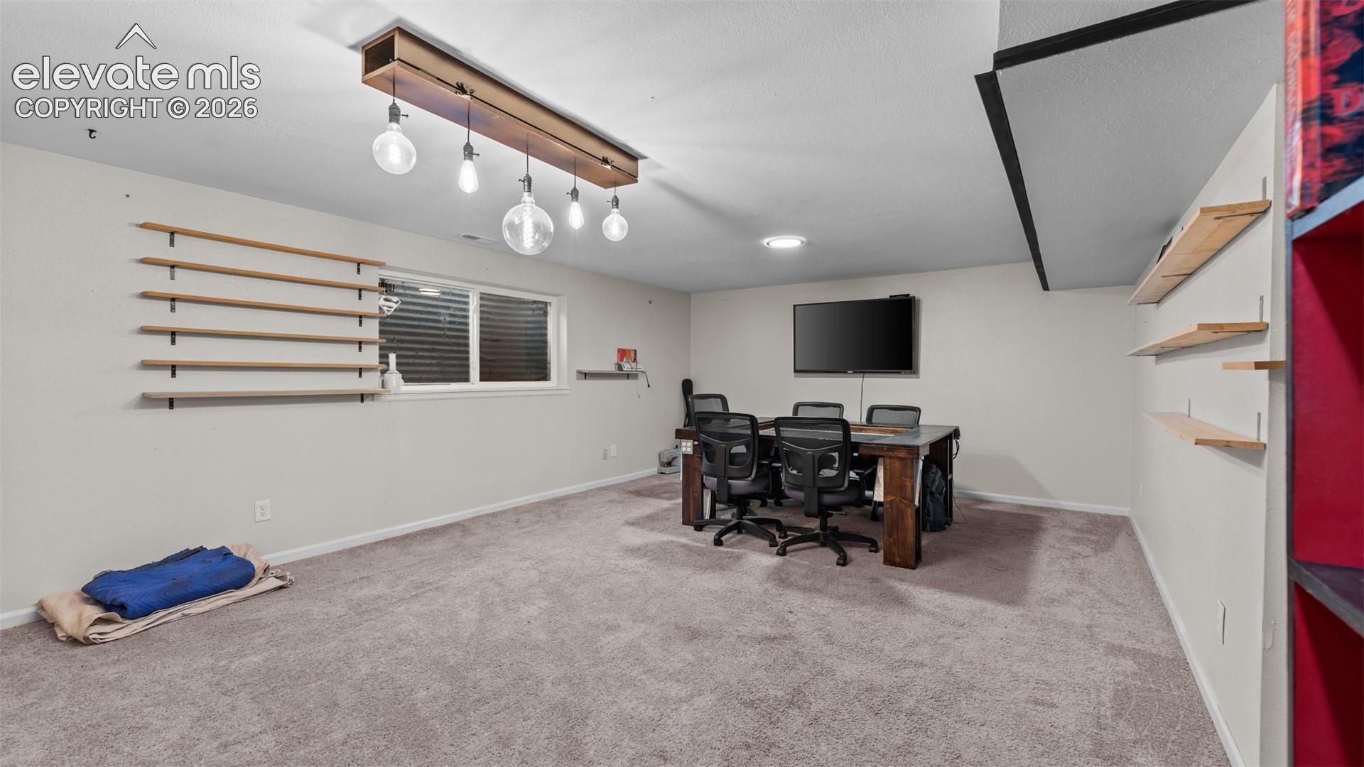 Neutral-toned room featuring wall-to-wall carpeting, custom tiered wooden wall shelving, and a linear pendant light fixture with exposed filament bulbs