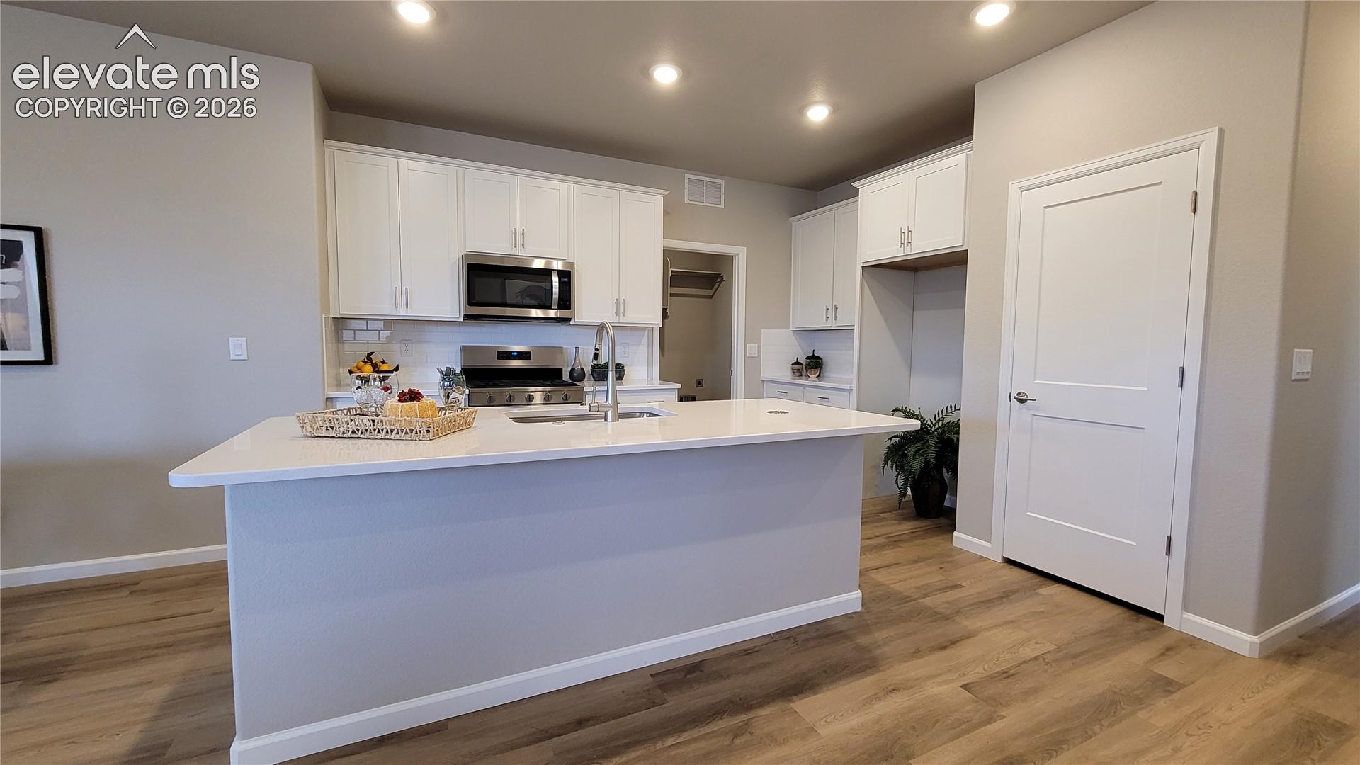 Kitchen featuring white shaker-style cabinetry, quartz countertops, a central island with a stainless steel sink, LED lighting, and LVP flooring, pantry with wood shelving, and stainless steel gas range, microwave, and dishwasher!