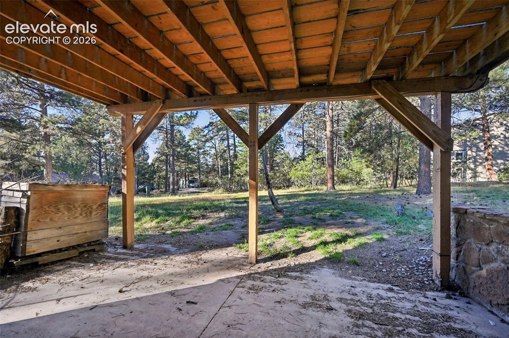 Covered patio space with concrete flooring, supported by exposed wood beams, set against a backdrop of mature trees