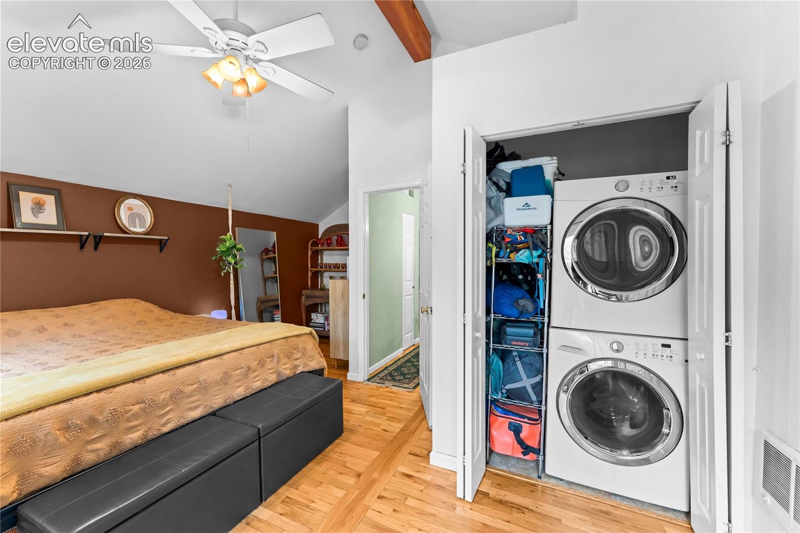 Vaulted ceiling with exposed wood beam, wood-finish flooring, ceiling fan with integrated light fixture, and built-in closet containing stacked laundry appliances