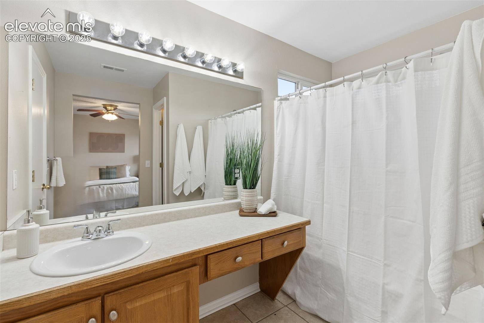 Vanity featuring a light-toned countertop, integrated oval sink, and wood cabinetry with multiple drawers, complemented by a large wall-mounted mirror, eight-bulb light fixture, and tiled flooring