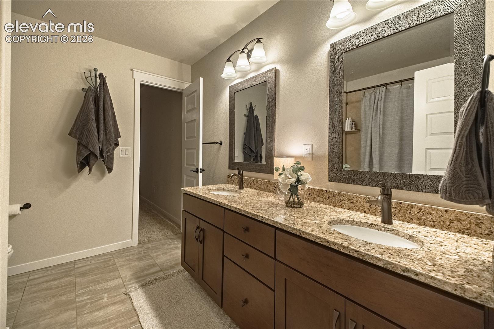 Dual-sink vanity with stone-finish countertop, undermount porcelain sinks, shaker-style wood cabinetry, framed wall mirrors, and multi-bulb vanity lighting