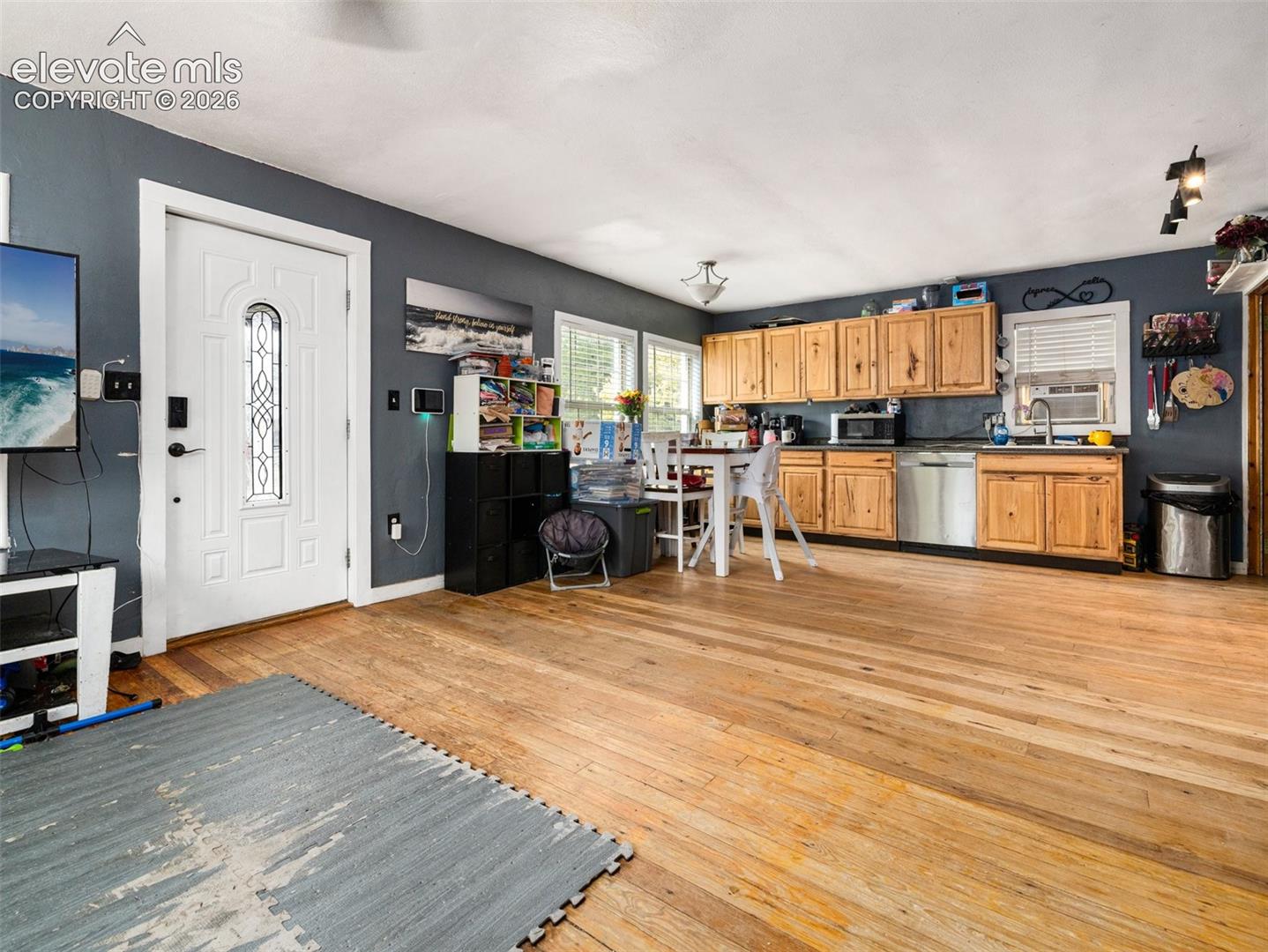 Natural wood-finish flooring throughout, kitchen with wood cabinetry, stainless steel dishwasher, and a white entry door with decorative glass paneling