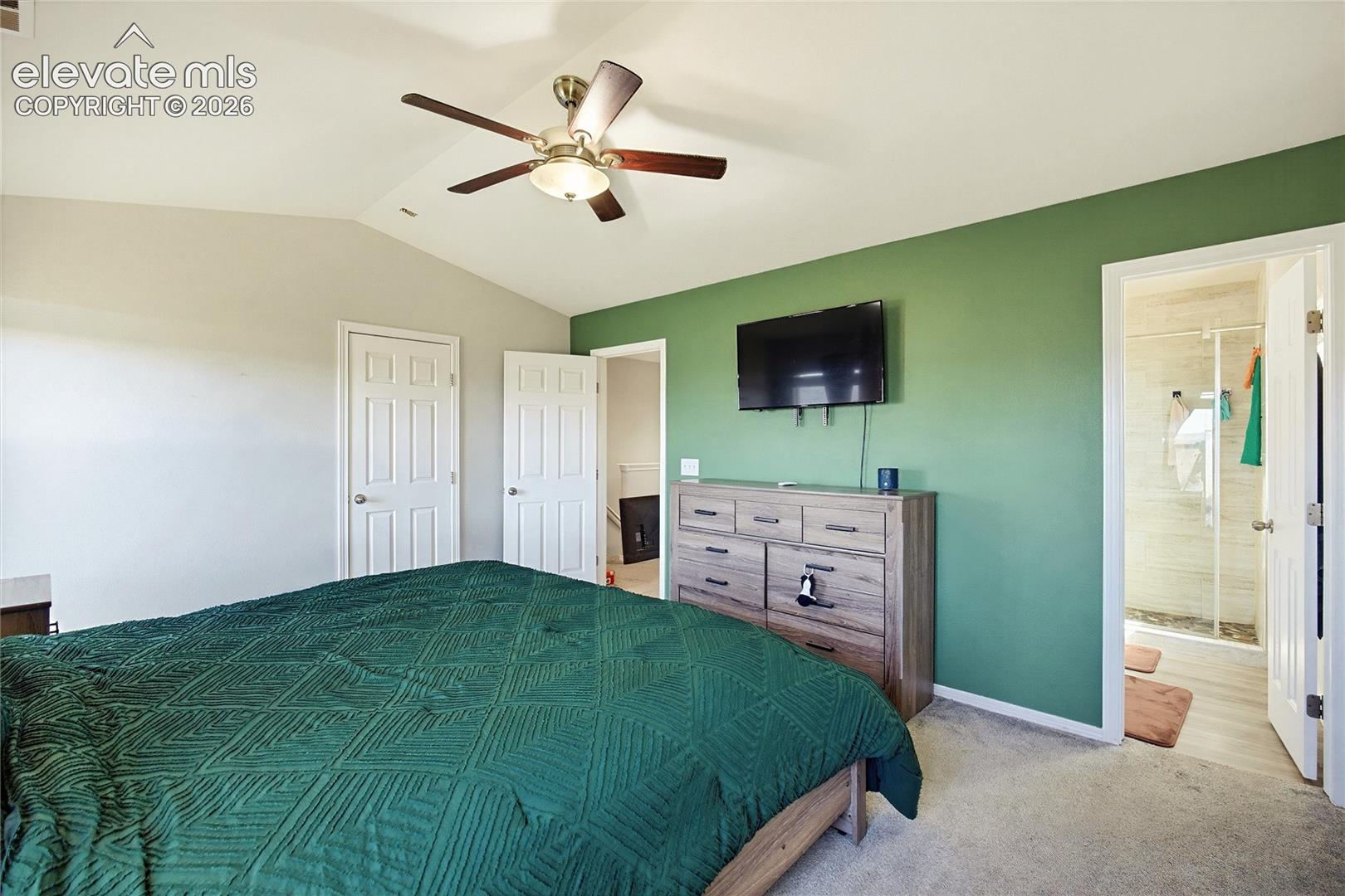 Vaulted ceiling, ceiling fan with light fixture, carpeted flooring, paneled doors, and an en-suite bathroom with a glass-enclosed shower