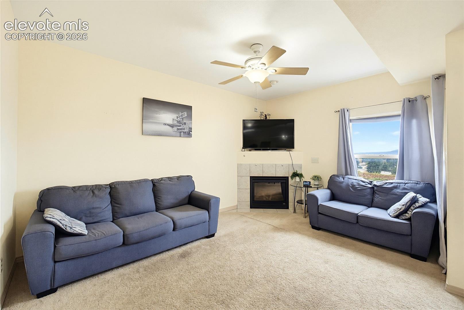 Living area featuring a corner fireplace with tile surround, a large window, ceiling fan with integrated lighting, and neutral carpeting