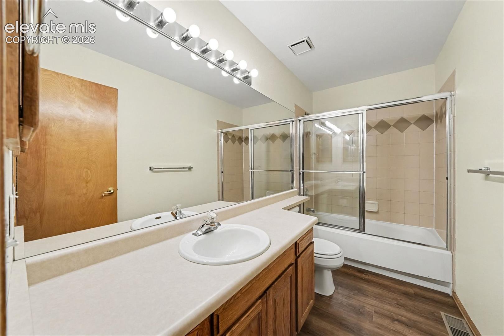 LVP flooring, full vanity with integrated sink, large wall-mounted mirror, and a tiled shower/tub combination with sliding glass doors