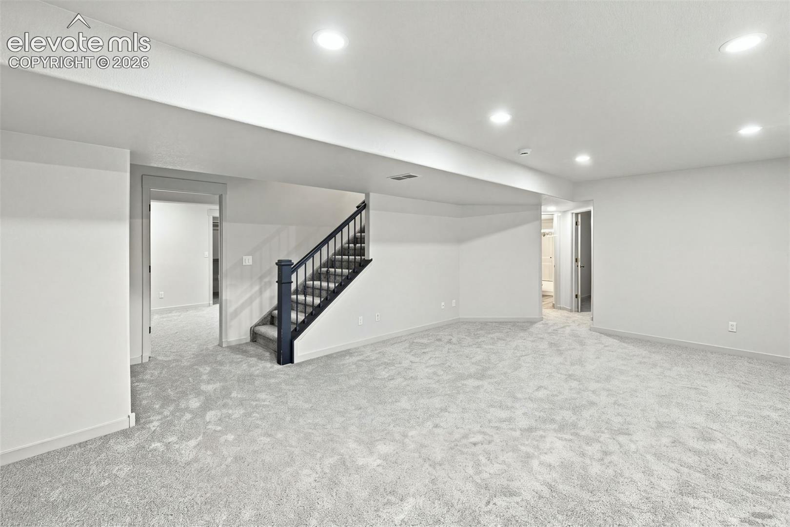 Open-concept interior featuring neutral carpeting, recessed ceiling lighting, and a staircase with a dark-finished handrail and balusters