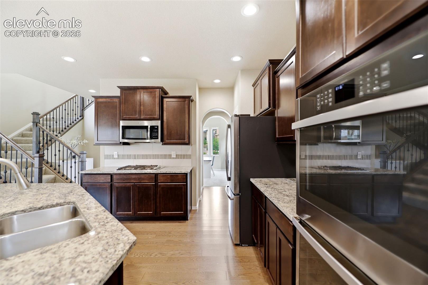 Kitchen featuring wood flooring, dark wood cabinetry, granite countertops, and stainless steel appliances including a double oven and refrigerator