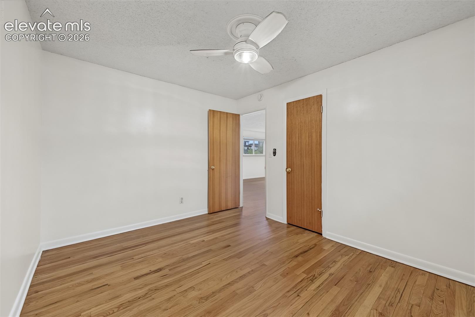 Room featuring wood-finish flooring, white walls, and a ceiling fan with integrated light fixture, punctuated by two wooden doors