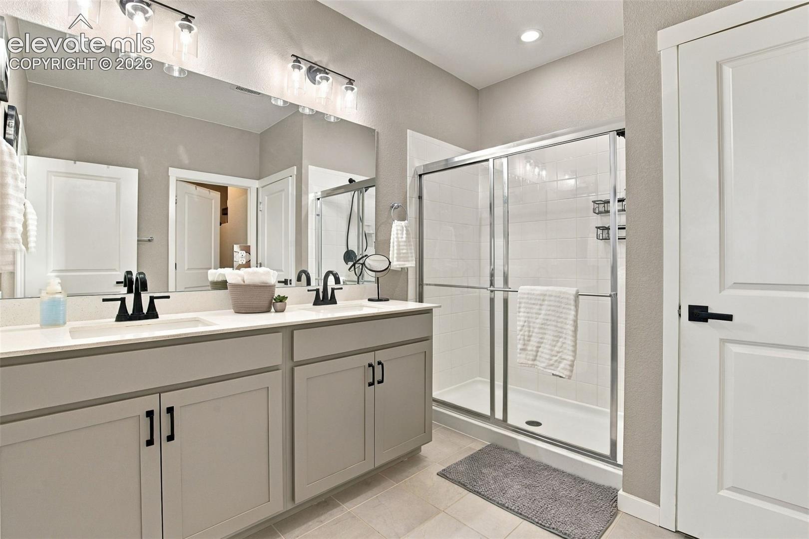 Dual vanity with neutral cabinetry, integrated double sinks, matte black fixtures, full-width vanity mirror with integrated lighting, and a walk-in shower featuring a sliding glass enclosure