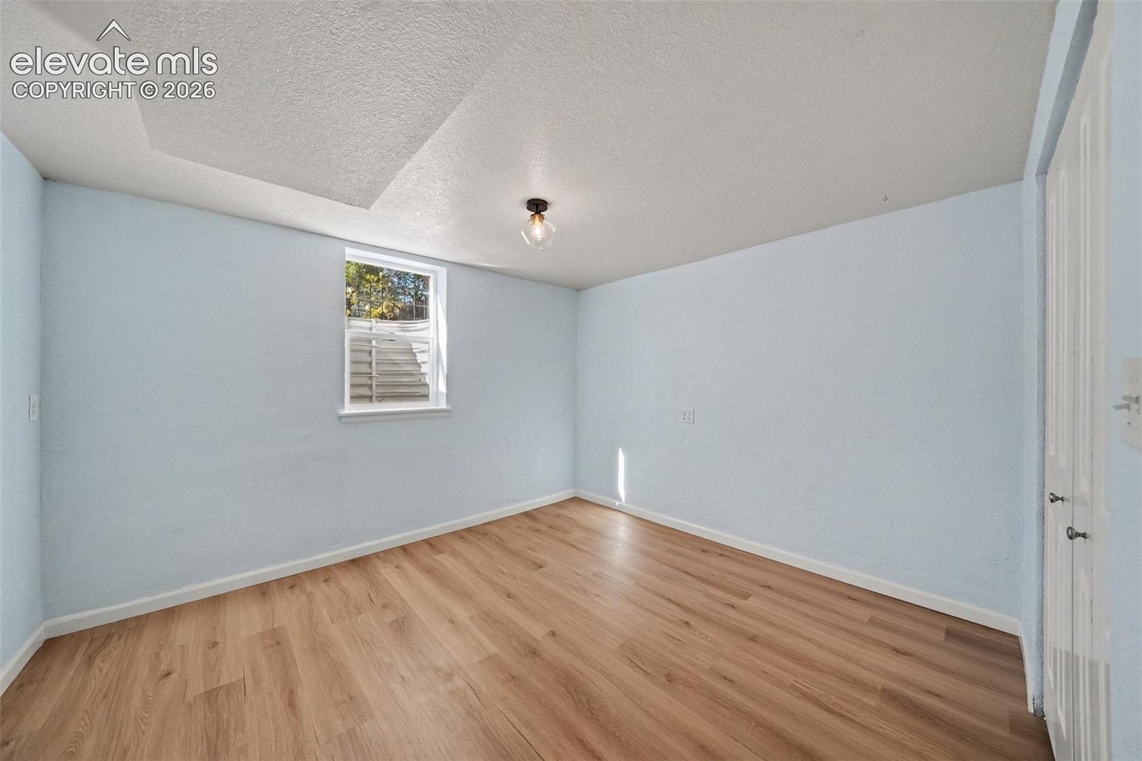 Room featuring wood-finish flooring, a recessed window with an egress well, a single-bulb flush-mount light fixture, and white dual-panel doors