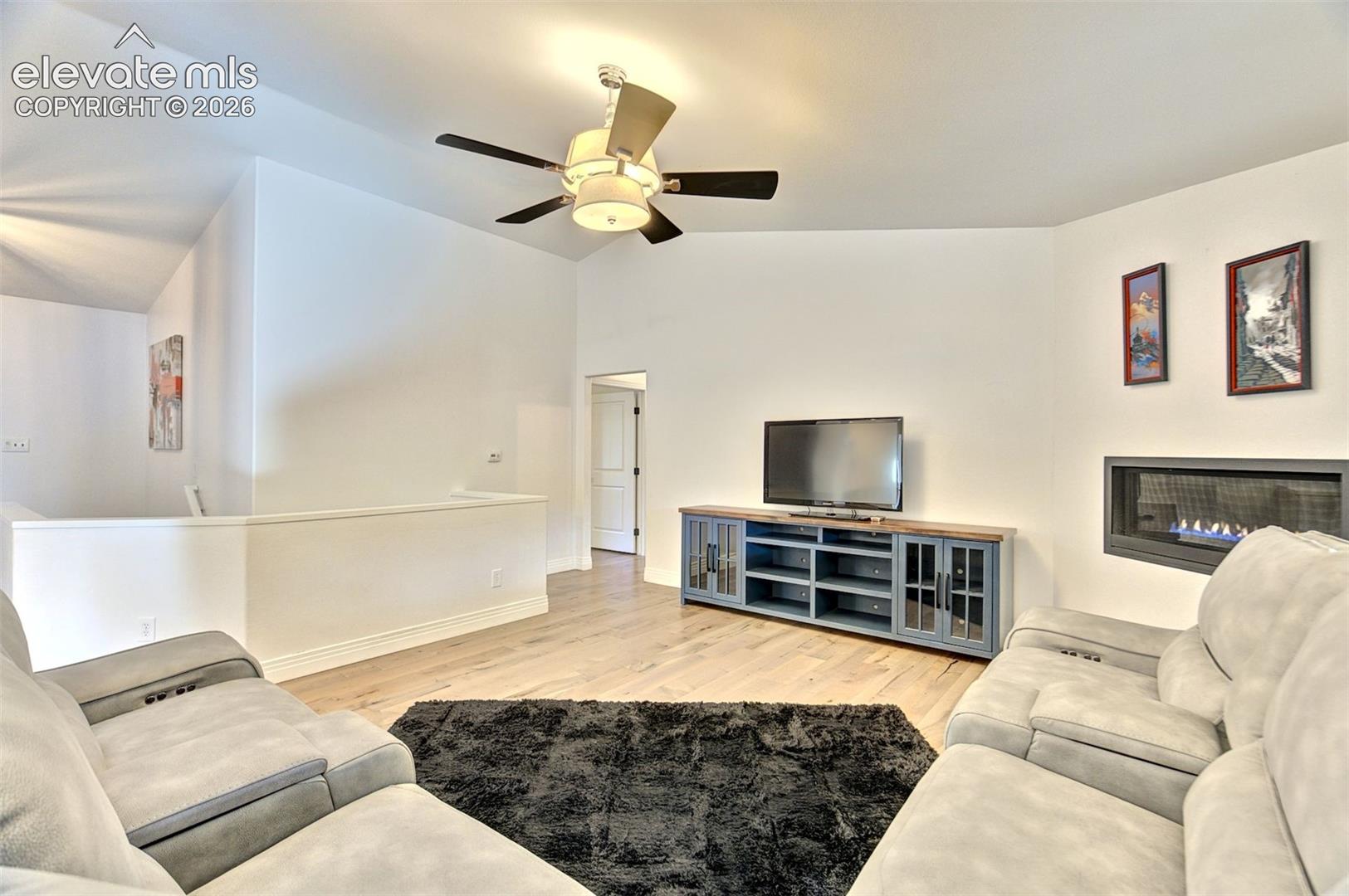 Vaulted ceilings, wood-finish flooring, linear wall-mounted fireplace, and ceiling fan with integrated lighting