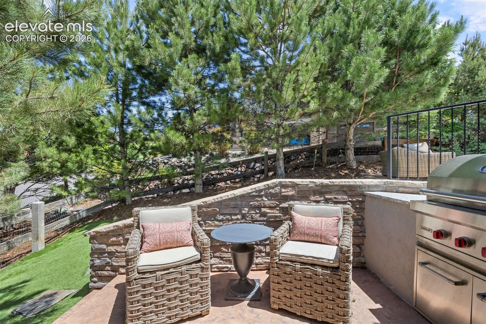 Paved patio featuring a built-in outdoor grill with stainless steel accents, a stone retaining wall, and mature pine trees