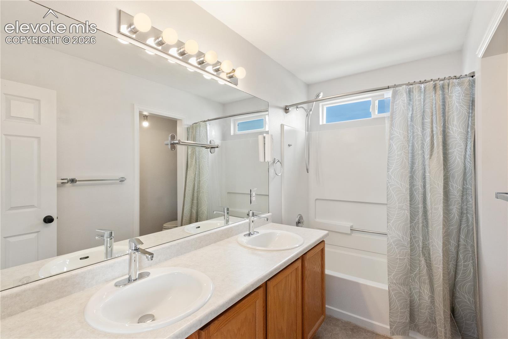 Dual-sink vanity with a solid-surface countertop and chrome fixtures, full-width vanity mirror, six-light wall-mounted fixture, and a tub-shower combination with an integrated surround