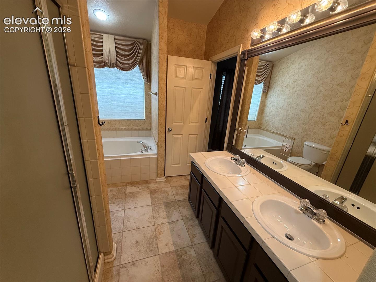 Dual-sink vanity with tile countertop, tile flooring, glass-enclosed shower, and deep soaking tub with tile surround