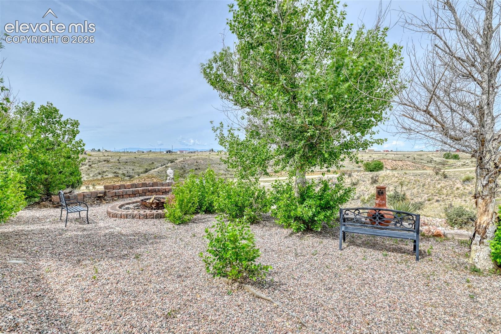 Outdoor entertaining area featuring a circular stone fire pit, gravel ground cover, mature trees, and natural desert landscaping beyond