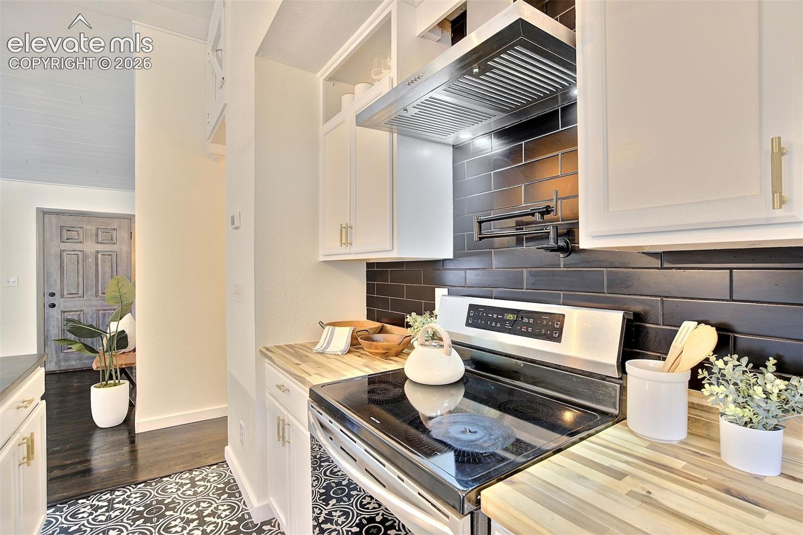 Kitchen featuring white shaker cabinetry, butcher block countertops, black subway tile backsplash, stainless steel range hood, and wall-mounted pot filler