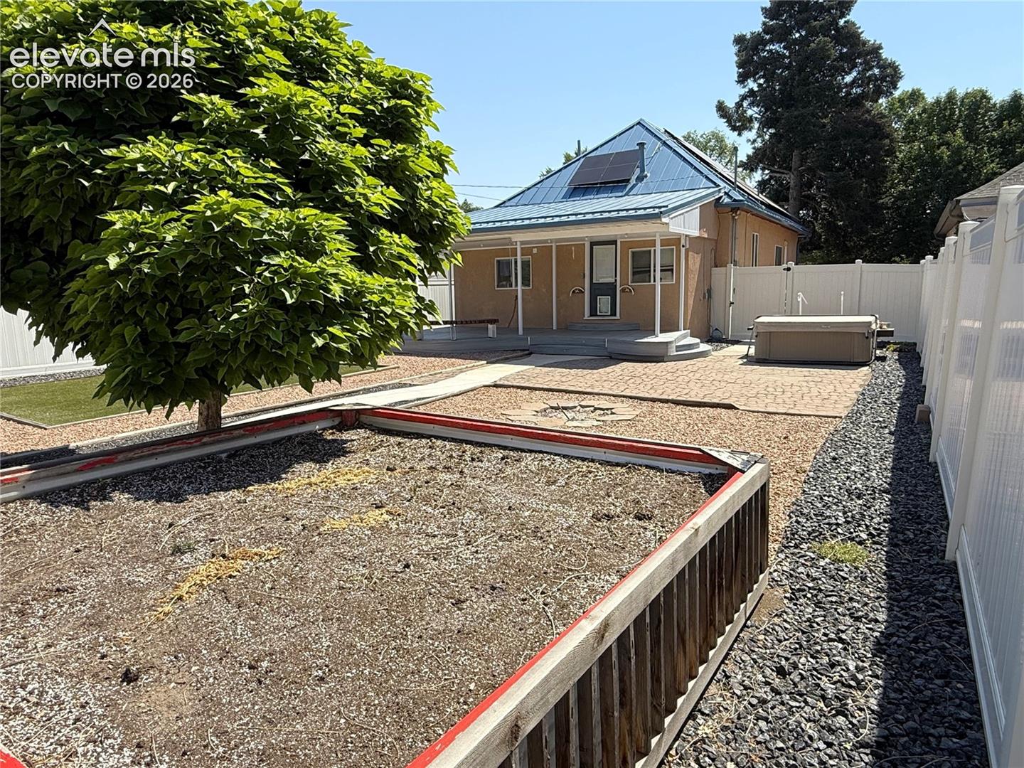 Exterior backyard featuring a metal roof, concrete patio, white vinyl fencing, raised garden bed with a wooden frame, and exterior solar panels