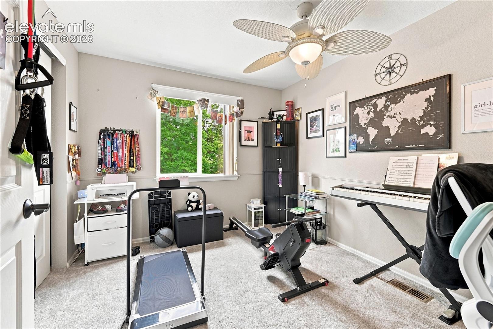 Neutral-toned interior space featuring light-colored wall-to-wall carpeting, a multi-blade ceiling fan with integrated lighting, and a window with exterior greenery