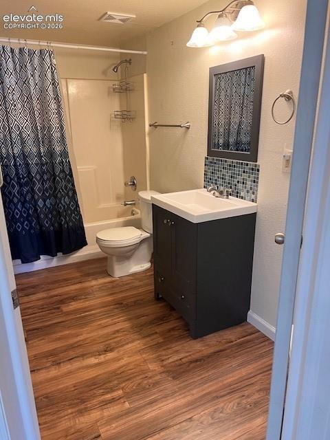 Bathroom featuring wood-finish flooring, a charcoal-toned vanity with a white basin, a glass-tile backsplash, and a three-light wall-mounted fixture
