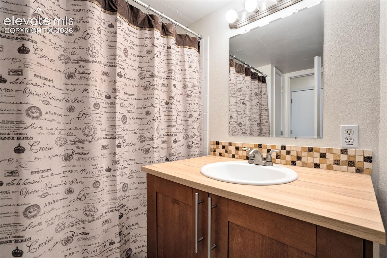 Bathroom vanity featuring wood-finish cabinetry, light-toned countertop, inset white basin, polished metallic faucet, and mosaic tile backsplash