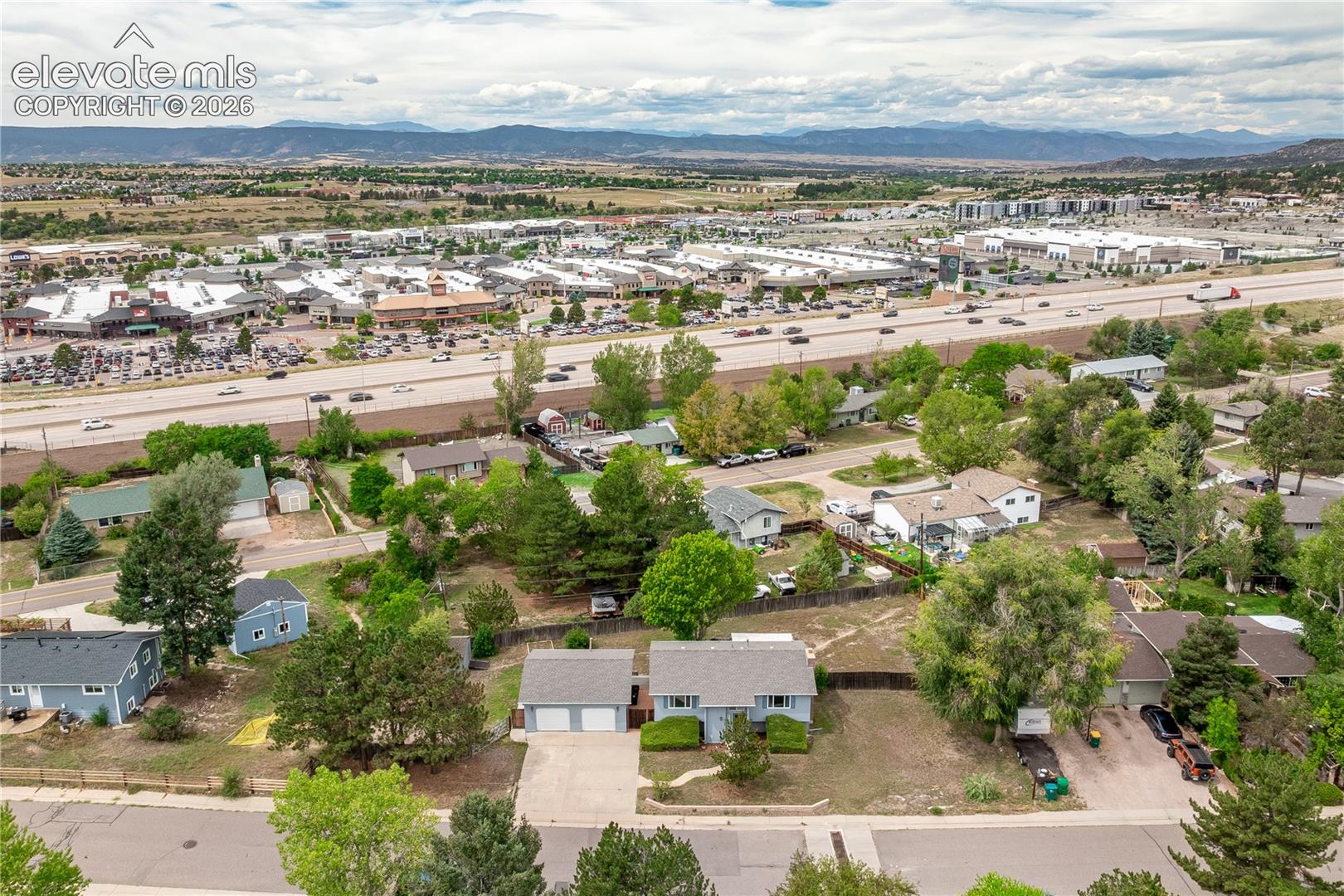 Aerial perspective featuring a single-story residence with an attached two-car garage, shingled roof, mountain backdrop, and established tree canopy