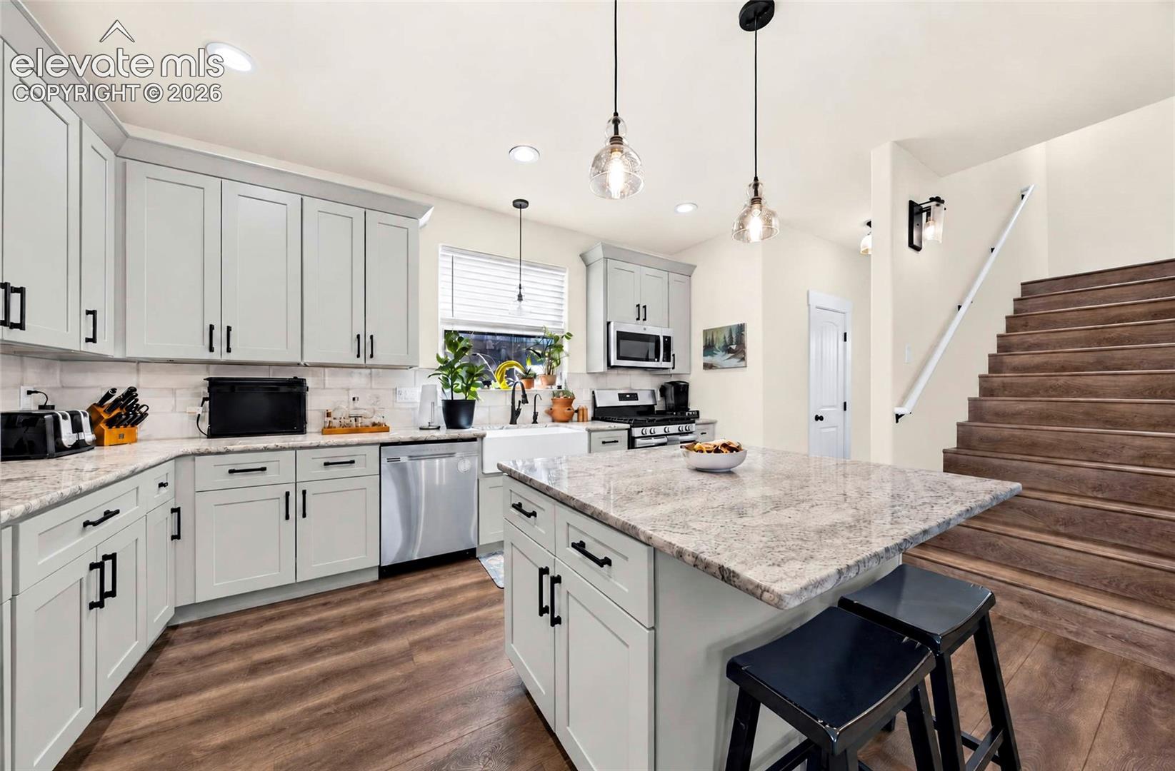 Kitchen featuring shaker-style cabinetry, stone countertops, a farmhouse-style apron sink, stainless steel appliances, and wood-finish flooring