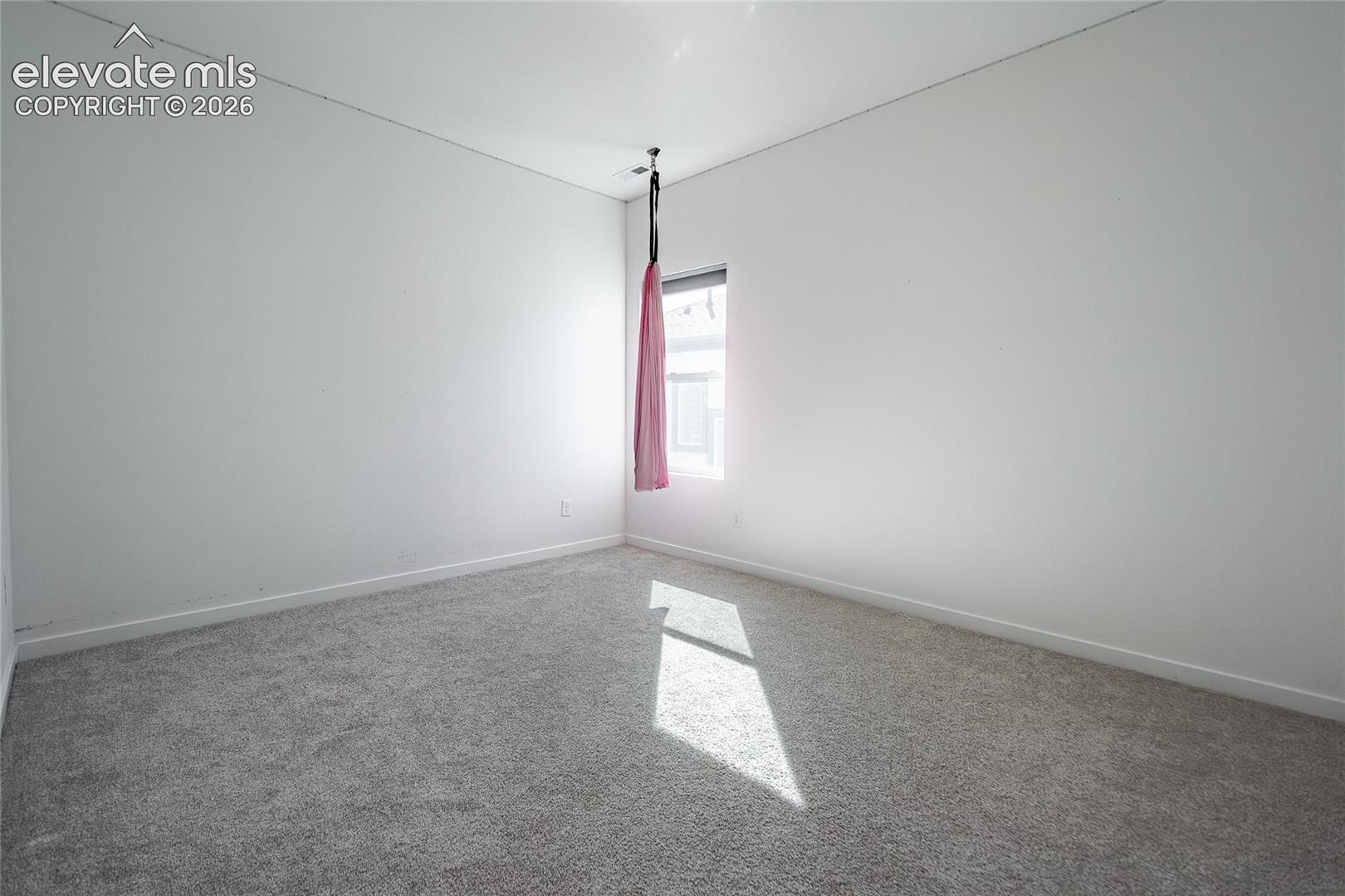 Neutral-toned interior space featuring light-colored wall paint, plush carpeting, a single window, and a ceiling-mounted hardware fixture