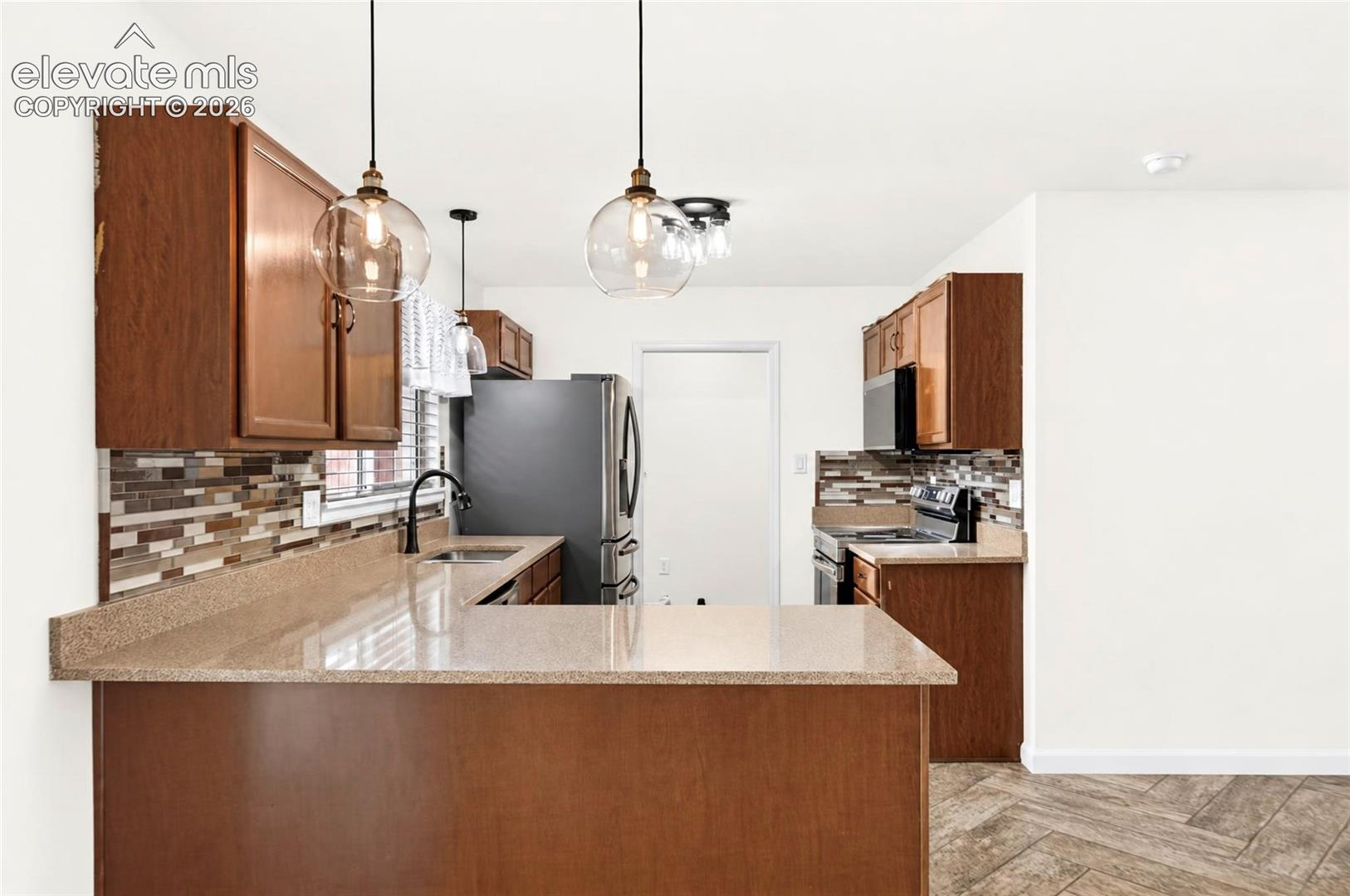 Kitchen featuring stained-wood cabinetry, stone-surface countertops, multi-toned tile backsplash, stainless steel appliances, and glass pendant lighting