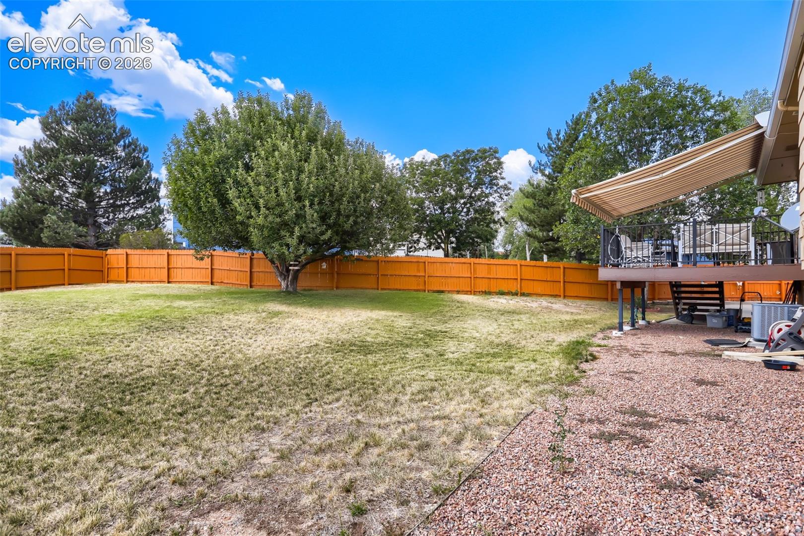 Expansive backyard featuring a wood privacy fence, mature trees, a raised deck with a retractable awning, and a gravel patio area