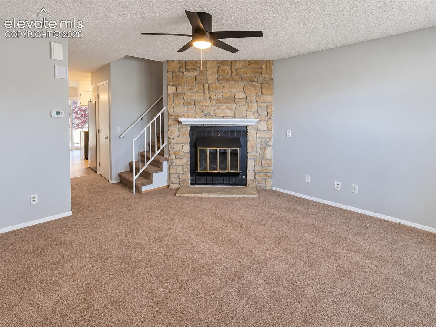 Floor-to-ceiling stone fireplace with a white mantel, black metal firebox, integrated ceiling fan, carpeted flooring, and staircase with a white metal railing