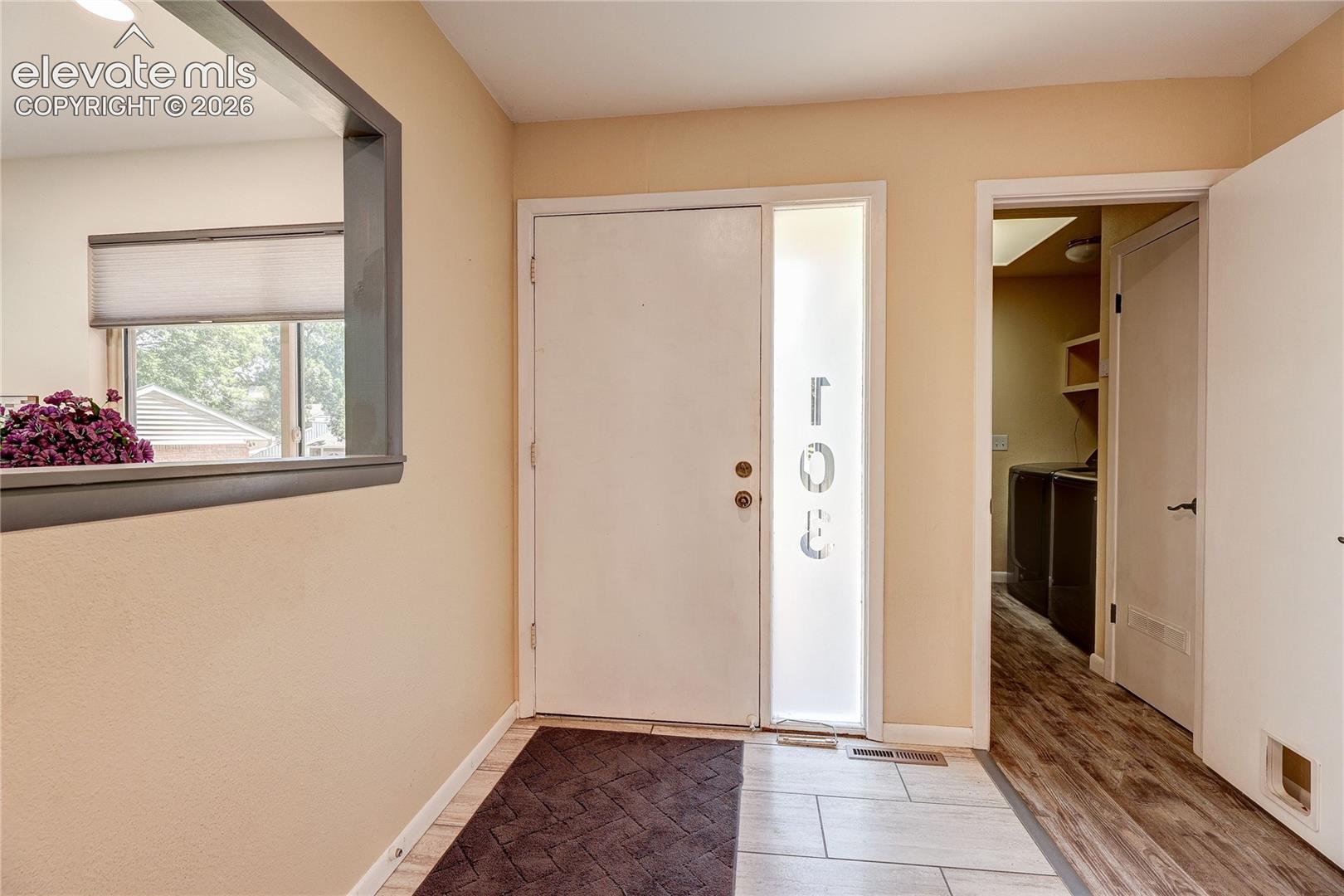 Entryway featuring a solid door with a vertical glass sidelight, interior window cutout with dark trim, tile flooring, and wood-finish flooring in the additional room