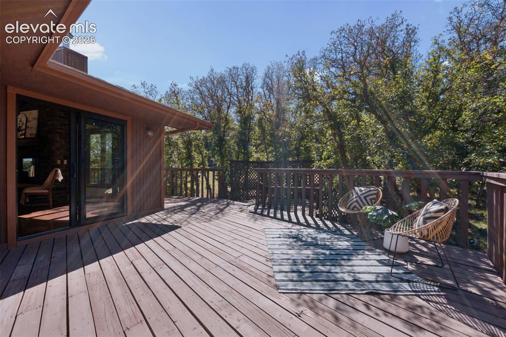Expansive wood-finish deck with a wood railing, surrounded by mature trees, and featuring a glass sliding door