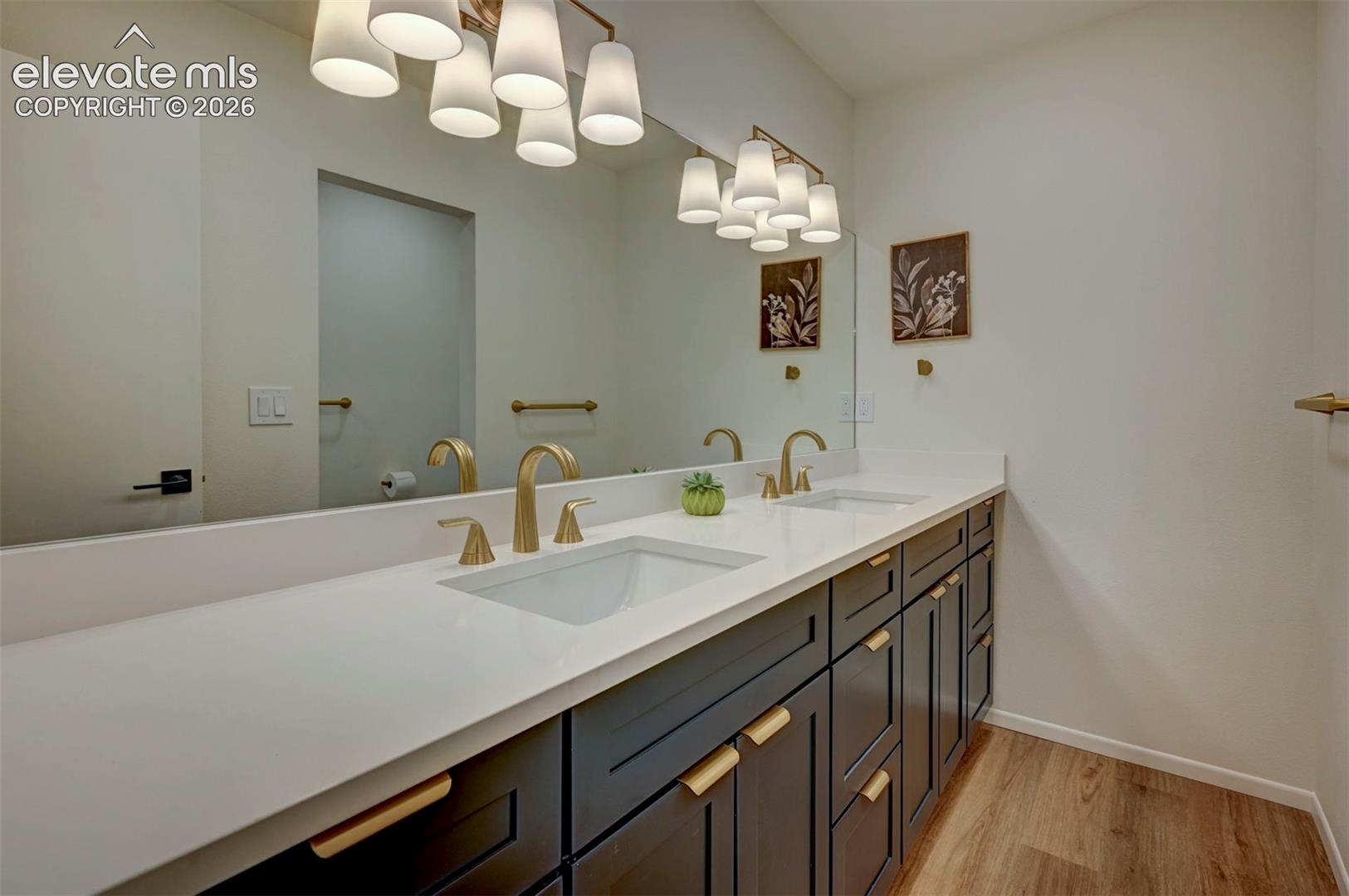 Double vanity featuring dark cabinetry, quartz countertops, dual undermount sinks with gold-toned fixtures, and a wide-format vanity mirror with multi-bulb overhead lighting