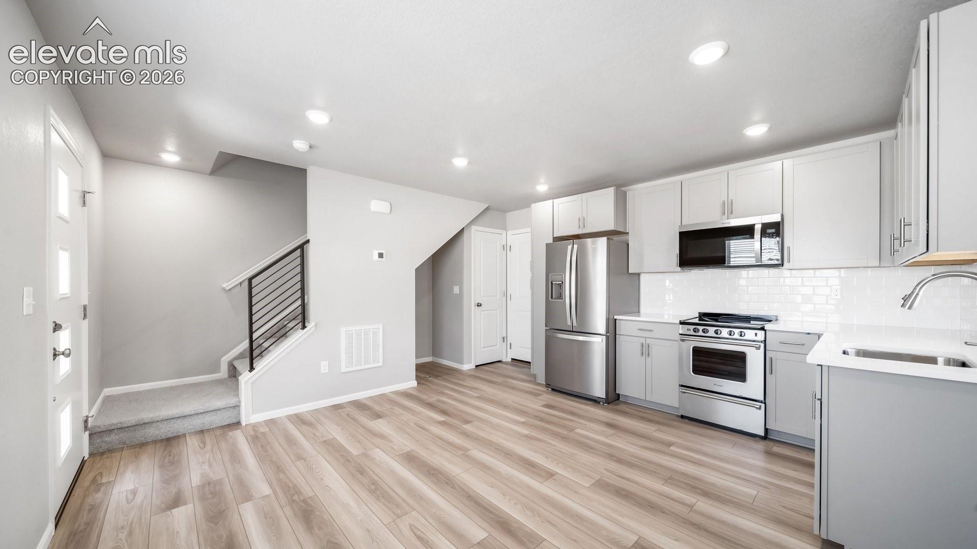 Kitchen featuring light-toned cabinetry, stainless steel appliances, subway tile backsplash, and wood-finish flooring, complemented by a staircase with matte black metal railings
*photos of a model home*