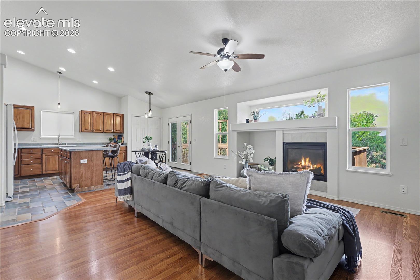 Open-concept living space featuring wood-finish flooring, a tiled kitchen area, a fireplace with a mantel, a ceiling fan, and recessed lighting