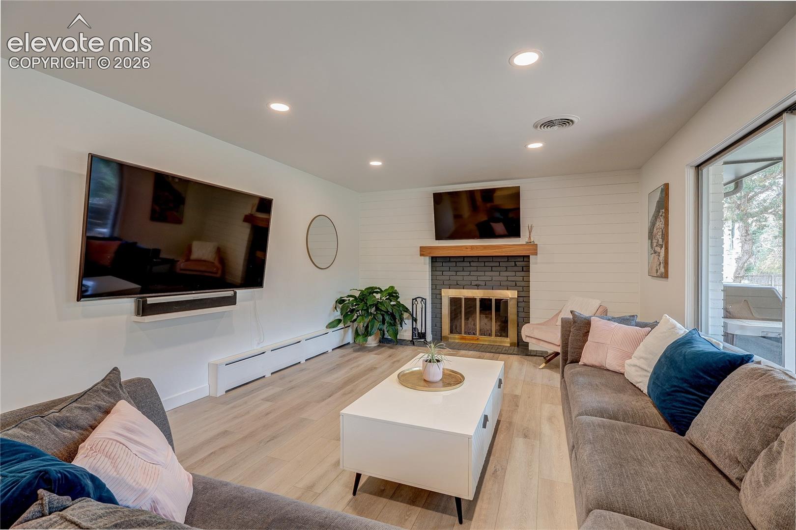 Spacious living area featuring wood-finish flooring, recessed lighting, and a fireplace with a brick surround and wood mantel