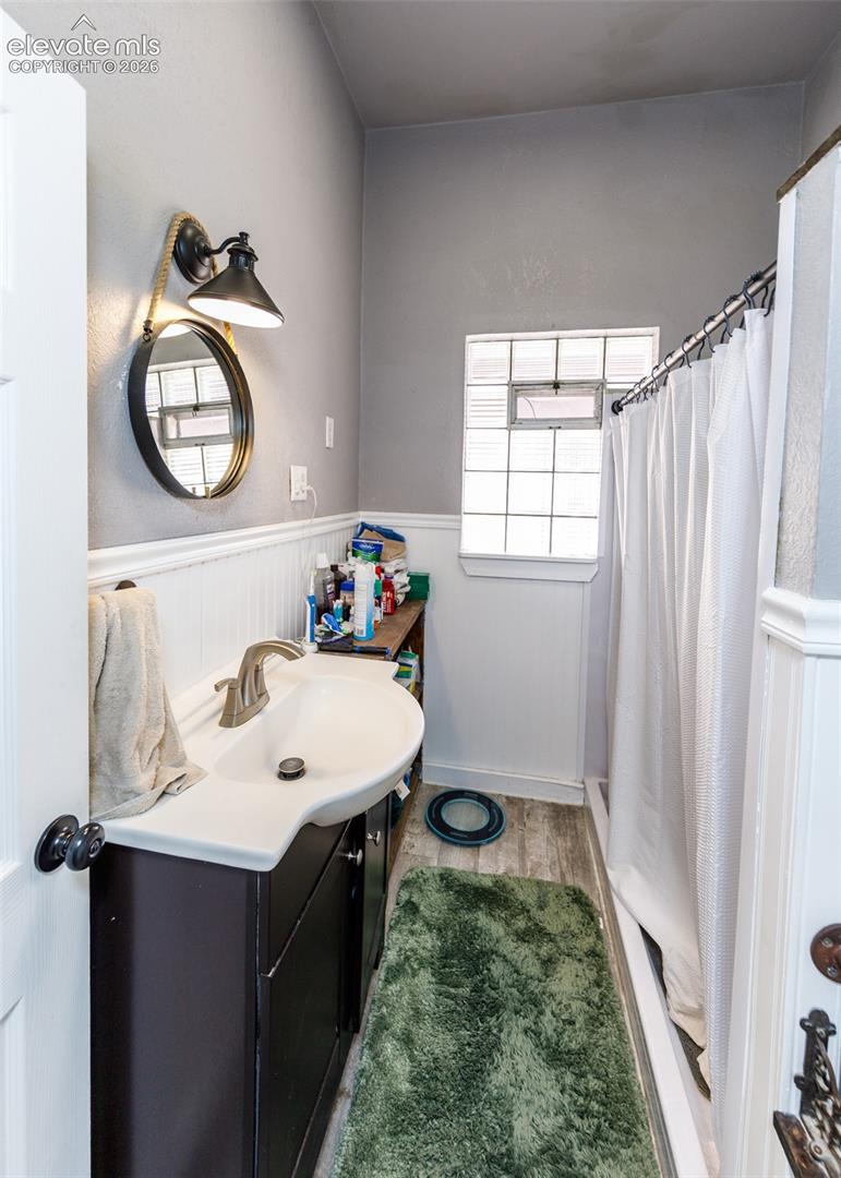 Vanity with integrated sink, wood-finish flooring, white wainscoting, and glass block window