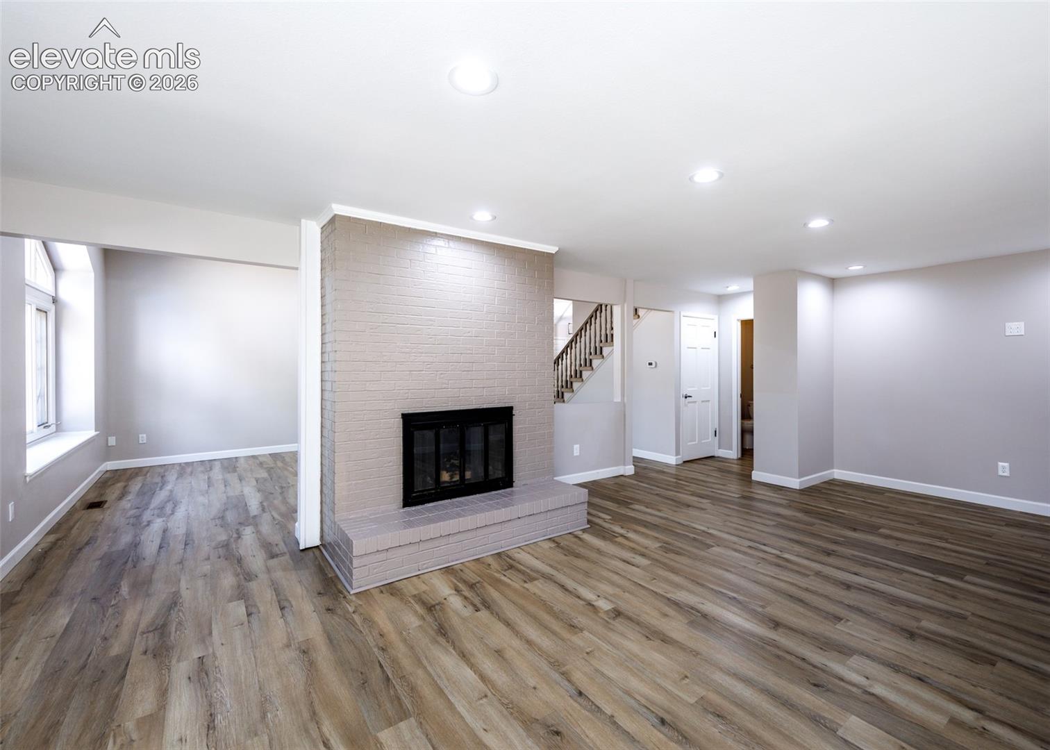 Expansive living area featuring wood-finish flooring, a painted brick fireplace, recessed lighting, and a staircase with wood banisters