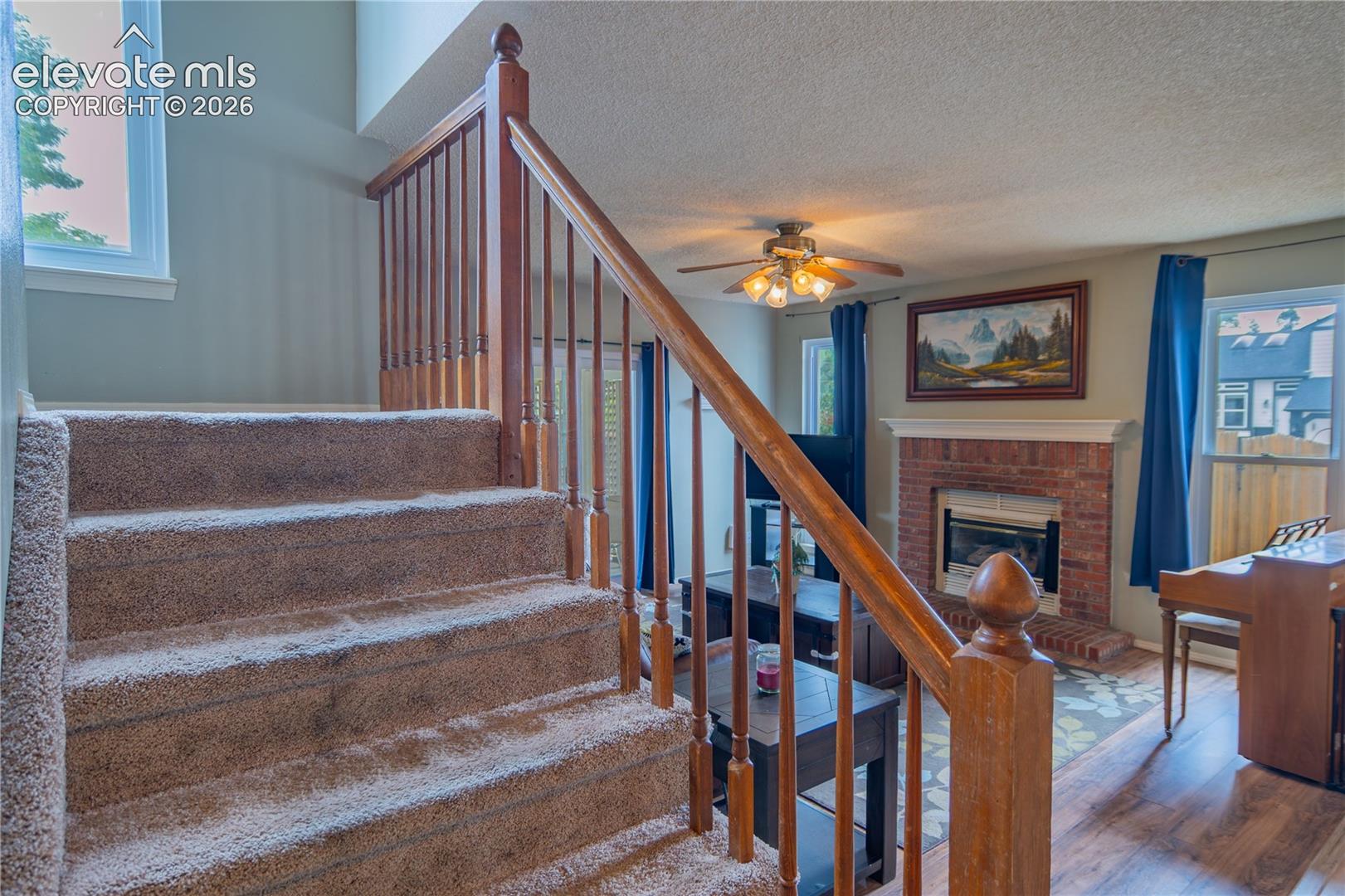 Carpeted staircase with wood railing, leading to a living area featuring a brick fireplace with a white mantel, wood-finish flooring, and a ceiling fan